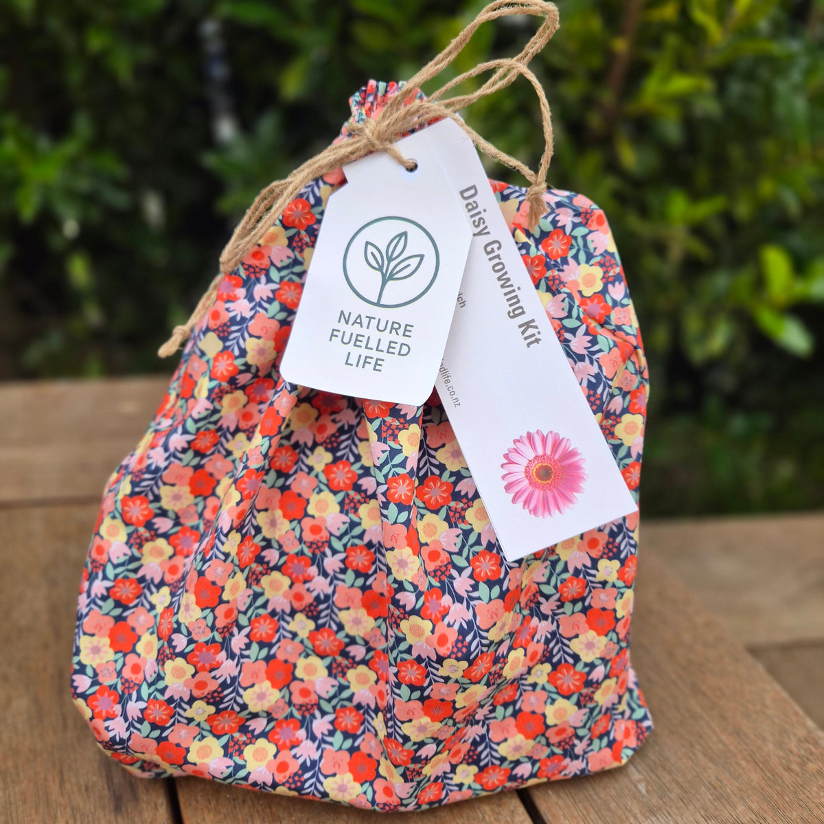 A small drawstring pouch with a dense floral print in orange, pink, yellow, and blue, sitting on a wooden table with a green garden background. It has a jute twine handle and two paper tags, one reading 'NATURE FUELLED LIFE' and the other 'Daily Glowing Kit'.